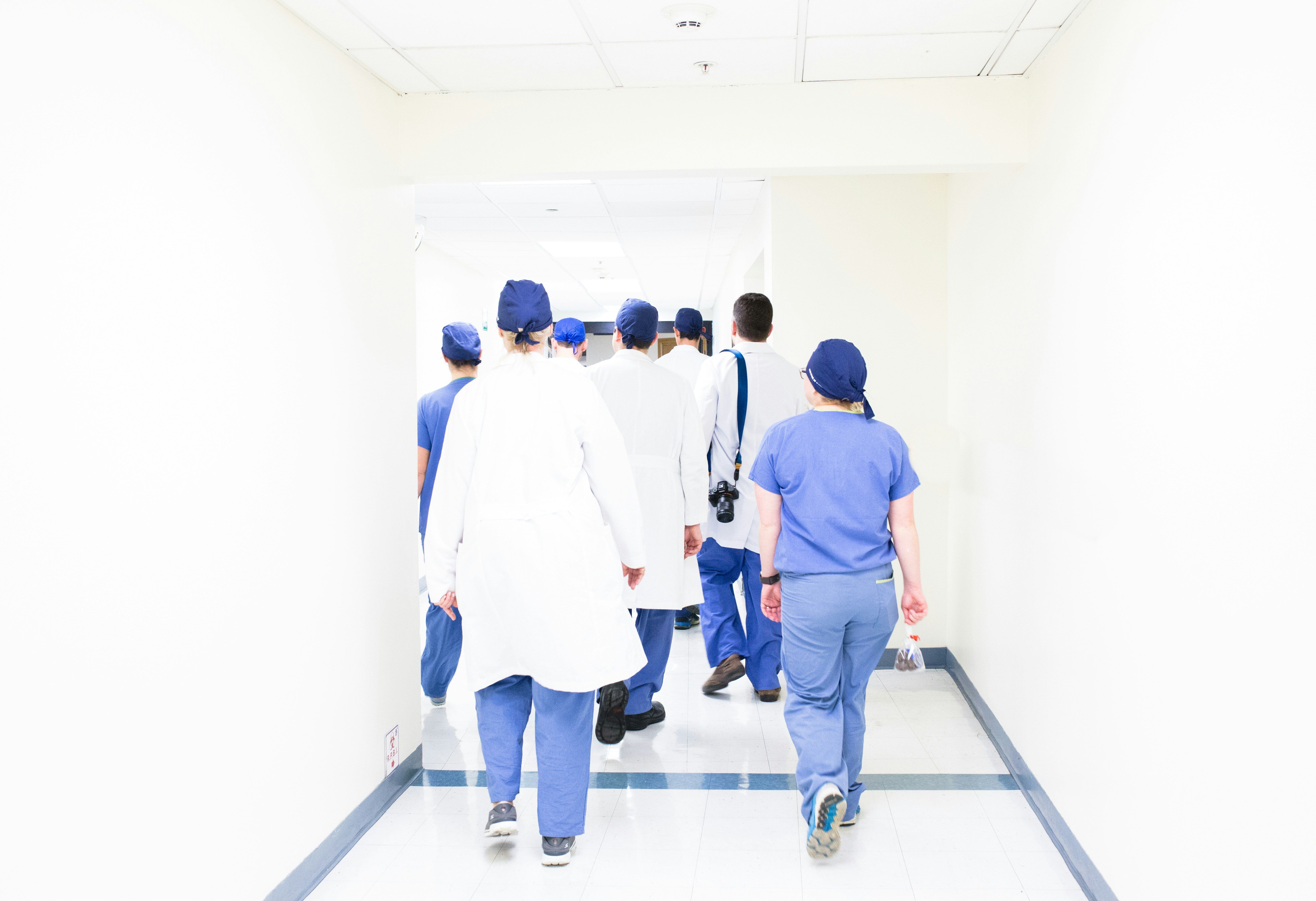 Une équipe de soignants en blouse et en tenue de bloc, vue de dos, avance dans un couloir d’hôpital baigné de lumière.